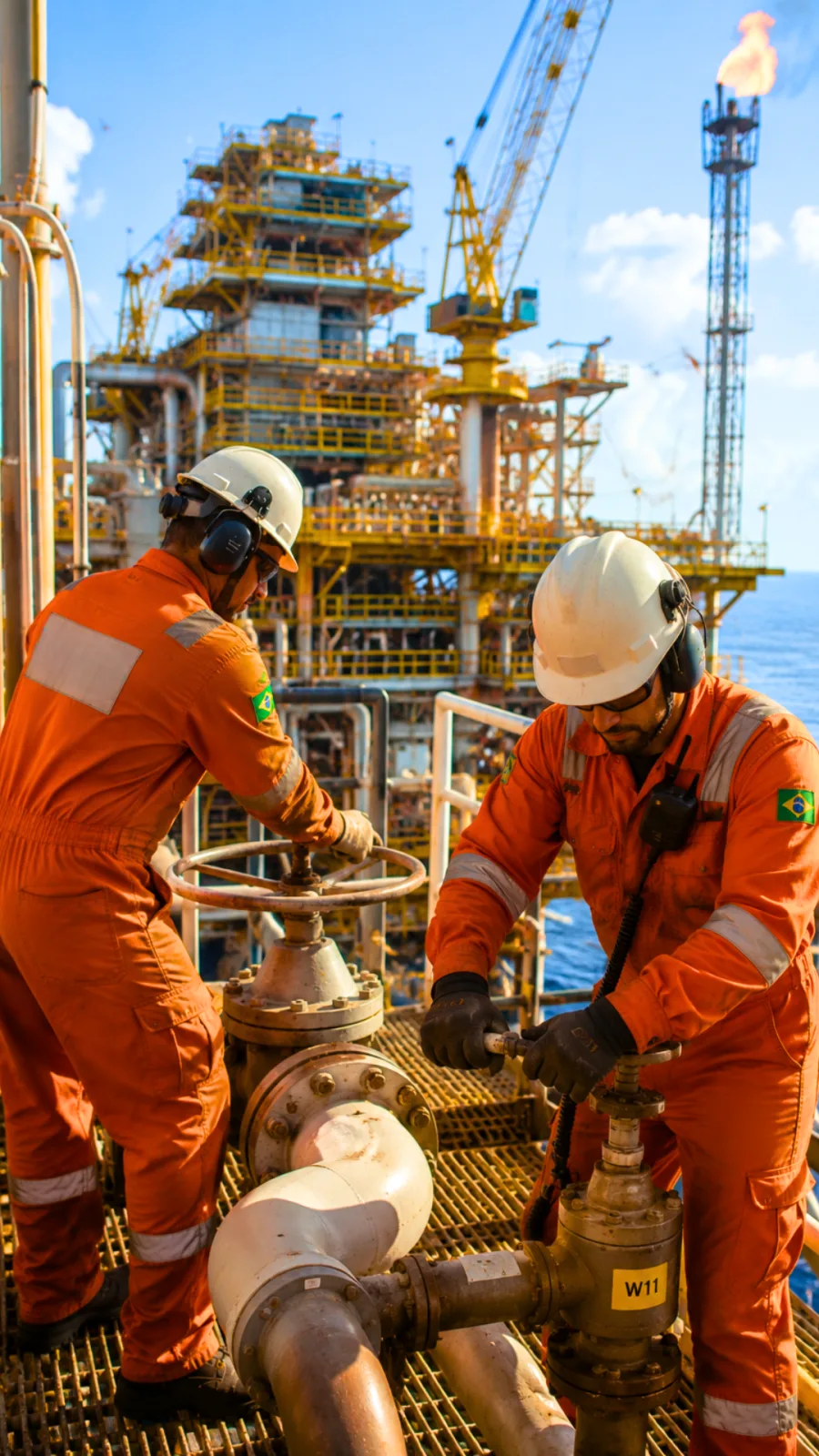 Dois operadores acionando válvula em plataforma offshore, com macacão retardante de chama laranja, faixas retrorrefletivas e capacete